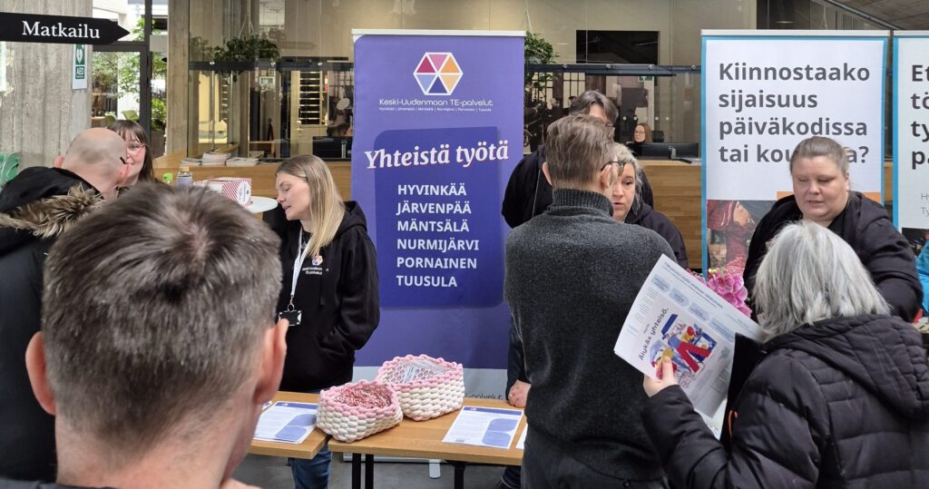 Työnhakijoita keskustelemassa Keski-Uudenmaan TE-palveluiden esittelypisteellä rekrytointitapahtumassa. Kuvassa useita ihmisiä tutustumassa työ- ja koulutusmahdollisuuksiin sekä vaihtamassa ajatuksia asiantuntijoiden kanssa sisätiloissa järjestetyssä tapahtumassa. Taustalla näkyy TE-palveluiden roll-up sekä rekrytointiin liittyviä infotauluja.